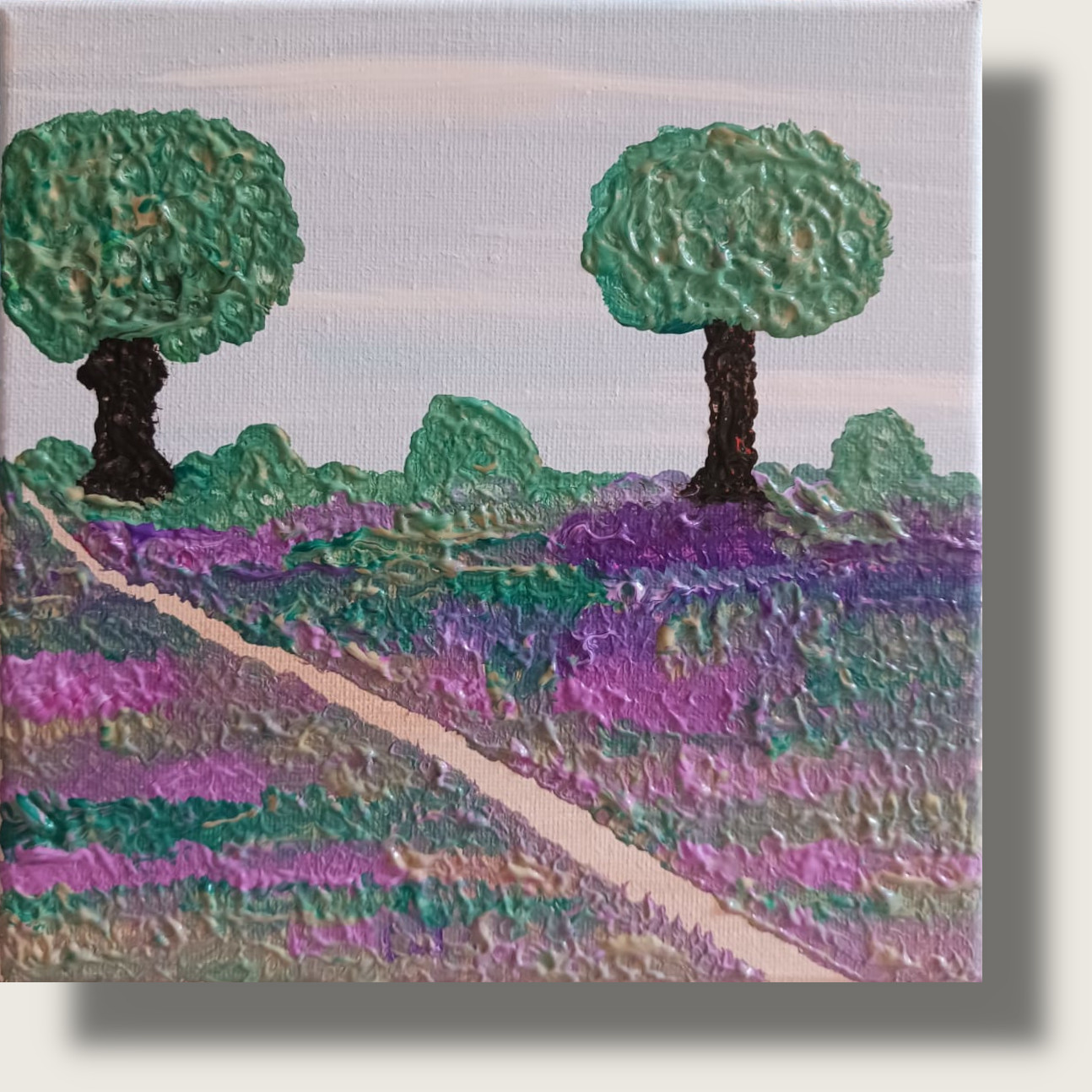 Lila Heidelandschaft der Maasdünen mit einem schräg führenden hellen Sandweg, grünen Büschen und zwei Bäumen vor blassem Himmel.